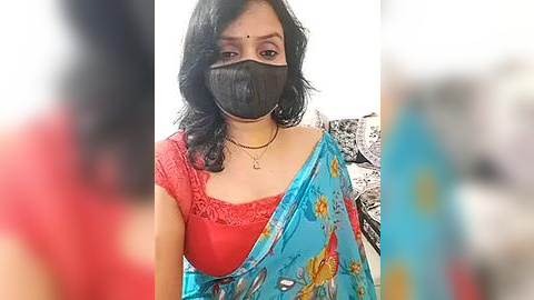 Media: Video of a woman with medium skin tone, wearing a black face mask, red lace top, and blue floral sari, standing against a blurred background.