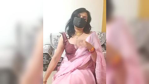 Media: Video of an Indian woman with medium skin tone, wearing a pink sari, black face mask, and black sunglasses, seated on a patterned couch in a living room.