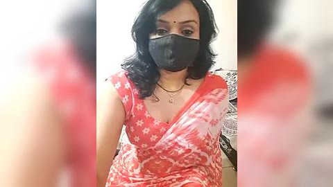 Media: Video of a South Asian woman with medium skin tone, wearing a black face mask, red tie-dye top, and black hair. Background is blurred.