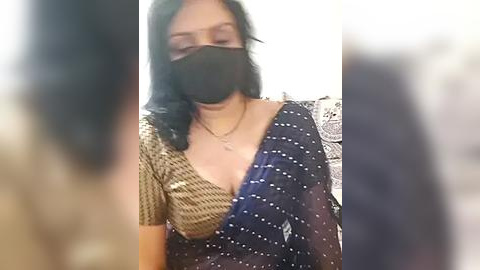 Media: Video of a woman in a black mask, dark blue polka-dot saree with a brown houndstooth pattern, wearing a necklace, indoors. Background blurred, showing a patterned wall.