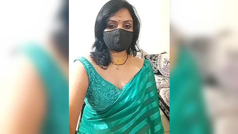 Media: Video of an Indian woman with medium skin tone, shoulder-length black hair, wearing a teal saree with black mask, sitting indoors with blurred background.