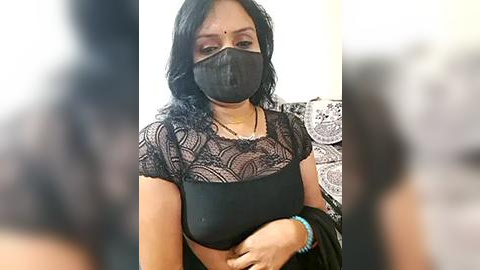 Media: Video of a woman with medium skin tone, long black hair, wearing a black lace top, black mask, and a blue bracelet. Background is blurred with intricate white lace patterns.