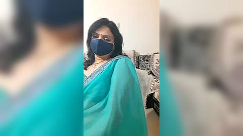 Media: Video of a middle-aged woman with dark skin, wearing a teal saree and blue face mask, standing in a blurred room with a white wall and framed art.