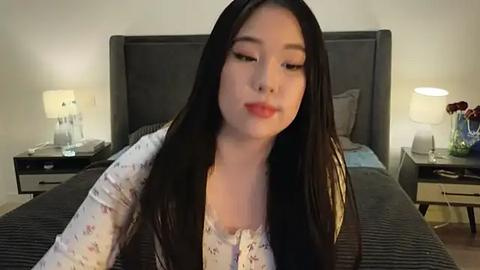 Media: Video of an East Asian woman with long black hair, fair skin, and red lipstick, wearing a white floral blouse, sitting on a bed with dark sheets, gray headboard, and two nightstands with lamps.
