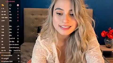Media: Video of a smiling, light-skinned woman with long, wavy blonde hair, wearing a white lace robe, seated on a bed with a beige headboard. Background includes a dark blue wall, a red flower vase, and a digital overlay showing social media metrics.