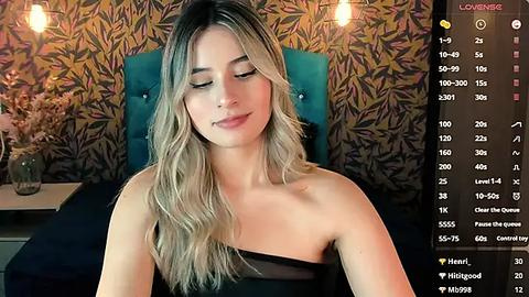 Media: Video of a young woman with wavy blonde hair, wearing a black strapless dress, seated in a room with a floral-patterned wallpaper and a teal chair.