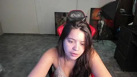 Media: Video of a young woman with long, dark hair in a red gaming chair, wearing a grey tank top, in a dimly lit room with grey carpet and black furniture.