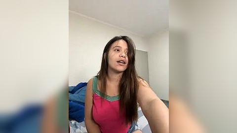 Media: Video of a young woman with long brown hair in a pink tank top, sitting on a bed with blue and white bedding in a simple, light-colored room.