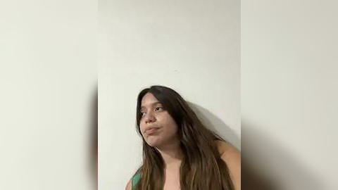 Media: Video of a young woman with long, straight brown hair, wearing a green top, standing against a plain white wall. She has a light skin tone and is gazing slightly upward, looking contemplative.