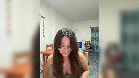 Media: Video of a young woman with long, dark hair, wearing a sleeveless top, sitting at a wooden table in a modestly furnished room with a tiled floor.