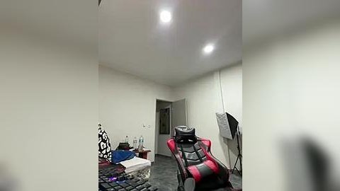 Media: Video of a dimly lit, cluttered bedroom with a red and black gaming chair, a patterned pillow, a water bottle, and a desk lamp, creating a messy yet functional gaming space.