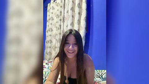 Media: Video of a smiling, light-skinned woman with long brown hair, wearing a black top, leaning on a bed with colorful patterned sheets. Background features a blue wall and white curtains with floral patterns.