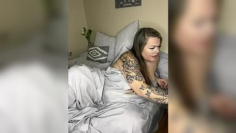 Media: A video of a tattooed woman with long brown hair, wearing a gray shirt, lying in a bed with gray sheets, surrounded by pillows and a vase with flowers, in a dimly lit room.