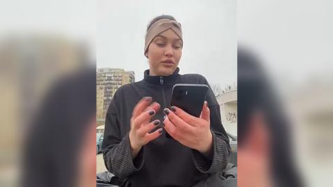 Media: A video of a woman in a black jacket and beige headband, using a smartphone, taken from a blurry perspective. The background shows an urban setting with buildings.