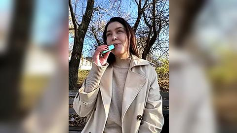 Media: Video of a woman in a beige trench coat, smiling while eating a blue popsicle outdoors, with leafless trees and a chain-link fence in the background.