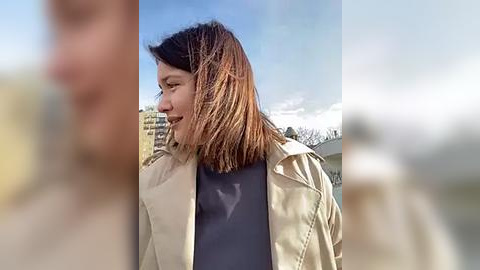 Media: Video of a Caucasian woman with straight, shoulder-length brown hair, wearing a beige trench coat over a dark top, standing outdoors in a city with blurred buildings and a clear blue sky.