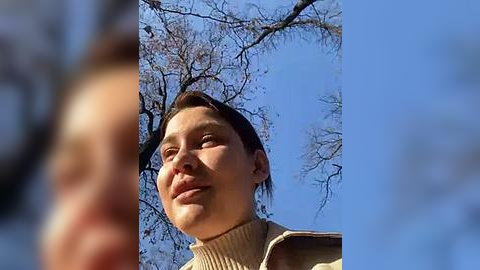 Media: Video of a young woman with short dark hair, smiling widely, wearing a beige turtleneck sweater, outdoors with a clear blue sky and bare tree branches in the background.