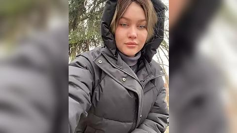 Media: Video of a young woman with light skin, wearing a dark grey puffer jacket, hood up, against a blurred outdoor background with greenery.