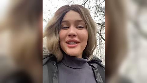 Media: Video of a young woman with shoulder-length, light brown hair, smiling widely, wearing a dark jacket and a high-necked sweater, set against a snowy, tree-filled background.