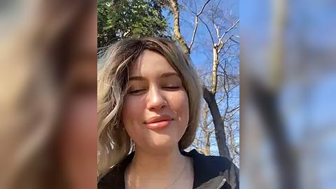 Media: Video of a young woman with light skin and shoulder-length blonde hair, wearing a black jacket, smiling and closing her eyes, standing outdoors with bare trees and a clear blue sky in the background.