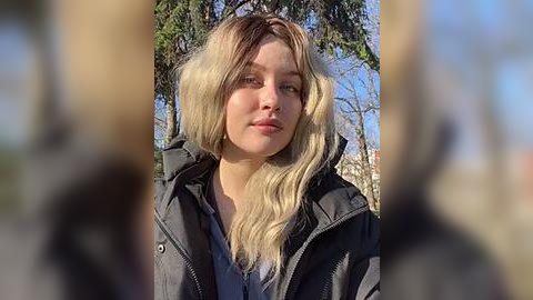 Media: A video of a young Caucasian woman with long, wavy blonde hair and fair skin, wearing a black jacket, standing outdoors with blurred background of trees and blue sky.