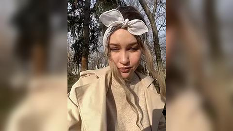 Media: Video of a young woman with fair skin, light brown hair tied with a white bandana, wearing a beige coat and a cream sweater, standing in a forest with blurred trees in the background.