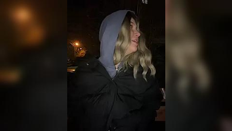 Media: A dimly-lit video of a blonde woman with wavy hair, wearing a hooded black jacket, and looking towards the left. The background features blurred figures and a warm, ambient light source.