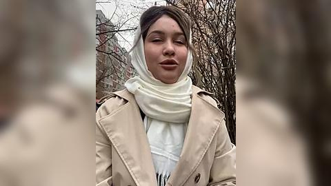 Media: Video of a young woman with fair skin, brown hair, wearing a beige trench coat, white scarf, and a white top, standing outdoors against a blurred background of bare trees and a brick building.