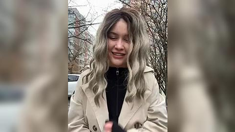 Media: Video of a smiling young woman with wavy, shoulder-length blonde hair, wearing a beige trench coat over a black turtleneck sweater, standing outdoors in a leafless urban environment.