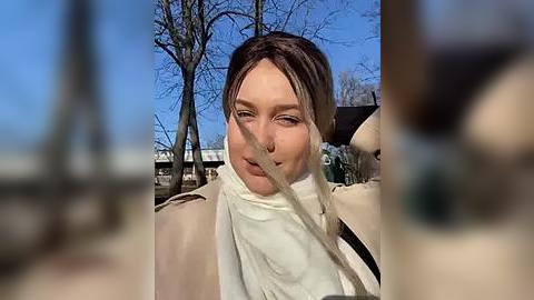 Media: A video of a Caucasian woman with short brown hair, wearing a beige coat, walking outdoors on a sunny day. The background features leafless trees and a blue sky.