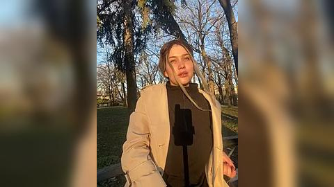 Media: Video of a young woman with fair skin and shoulder-length brown hair, wearing a beige coat and black turtleneck, standing in a sunlit park with bare trees and green grass.