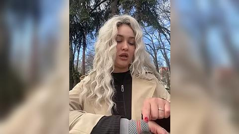 Media: A video of a young woman with long, wavy platinum blonde hair, wearing a beige jacket over a black top, standing in a forest with tall trees and a clear blue sky.