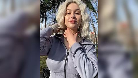 Media: Video of a blonde woman with wavy hair, wearing a light gray hoodie, standing outdoors, with blurred background featuring trees and a clear blue sky.