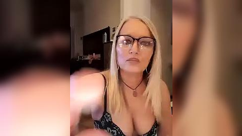 Media: Video of a blonde woman with glasses and a black floral-patterned bra, sitting indoors with blurred faces in the foreground.