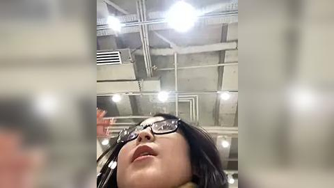 Media: Video of a woman with straight, shoulder-length black hair and black-framed glasses, looking upward. The ceiling features industrial lighting and exposed pipes. The image is slightly out of focus.