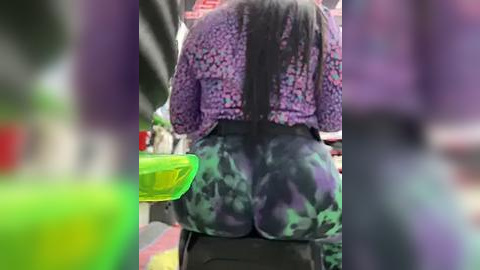 Media: Video of a woman in a purple floral shirt and tie-dye leggings, seated on a motorcycle with a green helmet in the background.