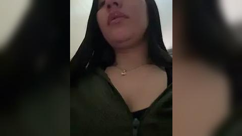 Media: Video of a woman with light skin, wearing a black hijab and a green top, featuring a small gold necklace. Her lips are slightly parted, and her face is partially obscured by the camera's angle.