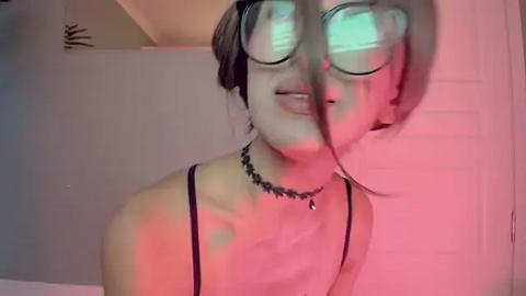 Media: Video of a young woman with pale skin and light brown hair wearing oversized, reflective glasses, a black choker, and a black spaghetti-strap top. She stands in a room with pink and green lighting, creating a surreal, dream-like atmosphere.