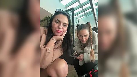 Media: Video: Two women, one with long black hair and sunglasses, and the other with braided blonde hair, sitting in a modern, glass-walled caf\u00e9, smiling, wearing winter coats.