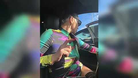 Media: A video of a person with short, dark hair and sunglasses, wearing a colorful, multicolored long-sleeve shirt, leaning out of a moving car, hand raised in a playful gesture.