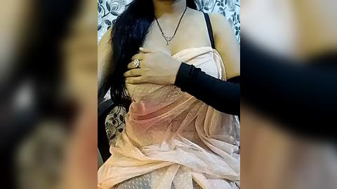 Media: A video shows a woman with long black hair in a sheer, pink chiffon dress, her breast exposed, being touched by a person's hand, wearing a black long-sleeve top, in a dimly lit room with a floral-patterned backdrop.