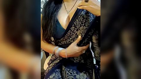 Media: A close-up video of a woman's chest in a dark blue, floral-patterned sari, revealing a black bra beneath. Her right hand rests on her left breast, emphasizing cleavage.