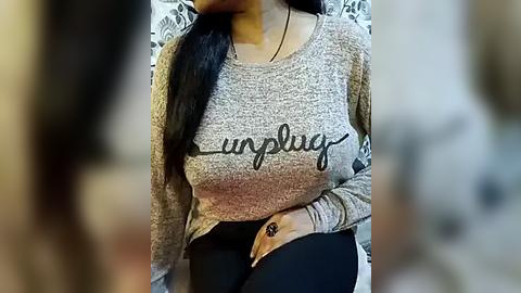 Media: Video of a woman with long black hair, wearing a gray, long-sleeve, \"sunflower\" t-shirt and black pants, sitting on a bed with floral patterned sheets, slightly out of focus.