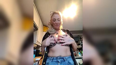 Media: Video of a blonde woman with medium build, wearing a leopard-print bra and denim shorts, holding her breasts, in a kitchen with beige cabinets, cluttered countertop, and bright light.