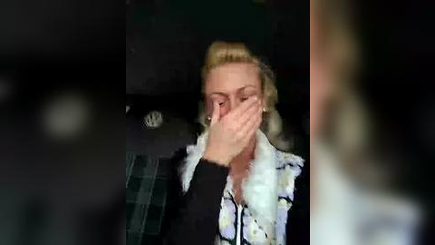 Media: Video of a blonde woman with a white fur collar, covering her face with her hands, in a dimly lit room with indistinct background.