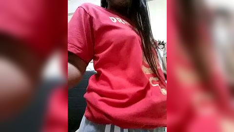 Media: Video of a woman in a red T-shirt with a white logo, wearing grey pants, standing indoors with blurred red and black background.