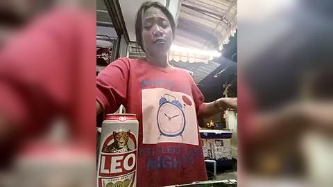 Media: A video of a middle-aged Asian man with short dark hair, wearing a red t-shirt with a clock graphic, standing in a dimly lit, cluttered store. He holds a red can of beer and a bottle of Leo beer.