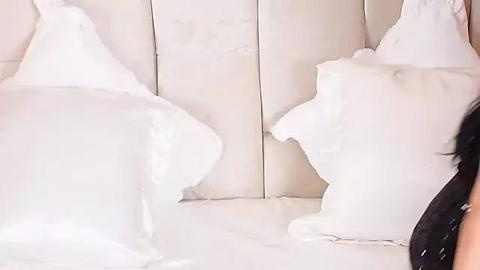 Media: Video of two white pillows in a plush, cream-colored bed, with a person's head and shoulder visible in the background, wearing a black top.