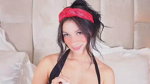 Media: Video of a young, fair-skinned woman with long, dark hair tied in a ponytail, wearing a red headband and black halter top, sitting on a white bed with beige padded headboard.