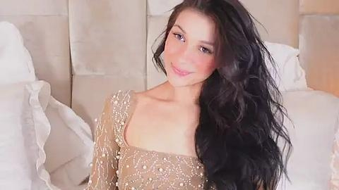 Media: Video of a smiling young woman with long, wavy black hair, wearing a sequined, long-sleeved, off-the-shoulder top, seated on a white bed with cream-colored pillows in a softly lit bedroom.